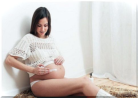 pregnant woman sitting next to wall puts cream on baby bump